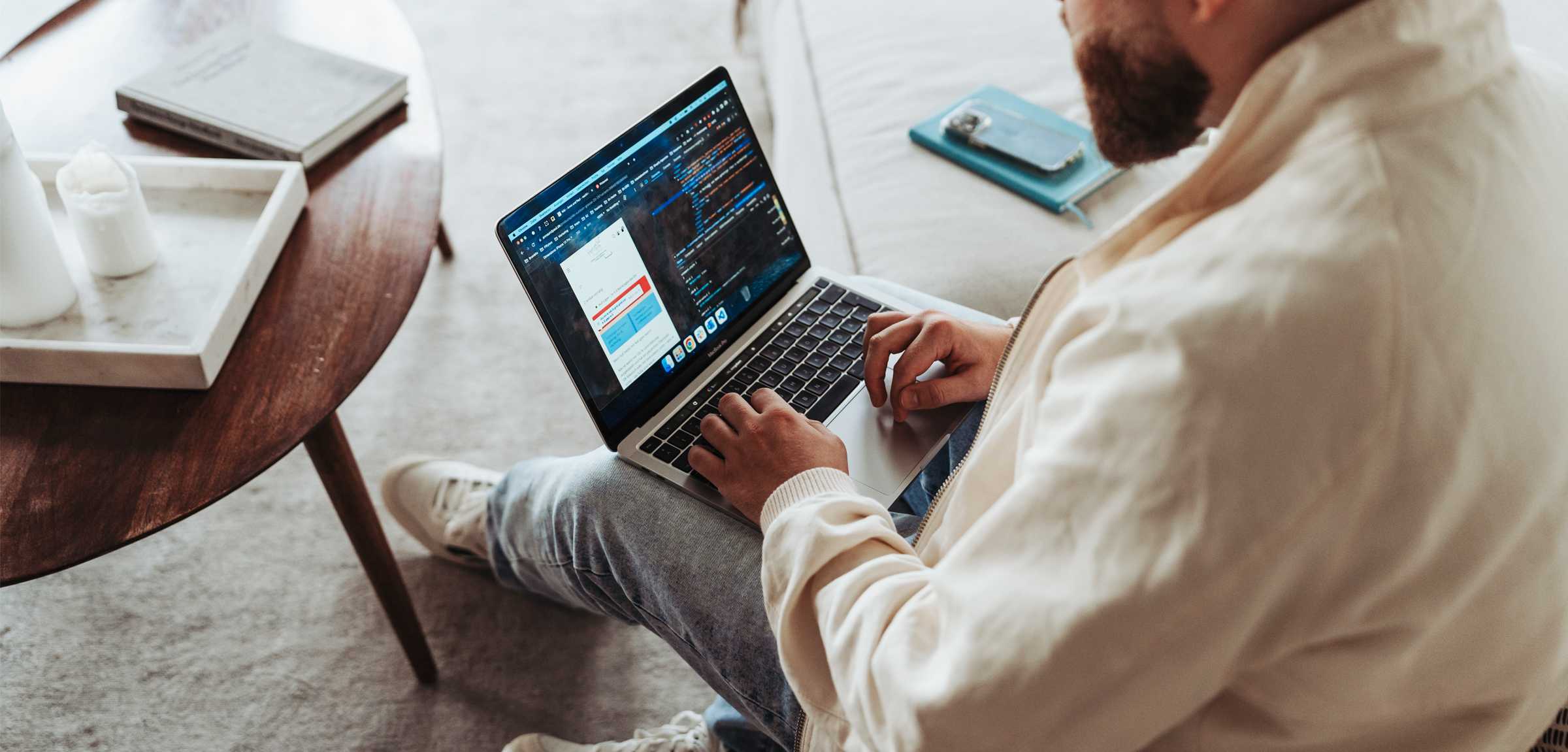 Ein Mann sitzt auf einem Sofa und arbeitet an einem Laptop. Auf dem Laptop ist ein Online-Shop zu sehen. Links steht ein Tisch, rechts auf dem Sofa liegen ein Buch und ein Smartphone.
